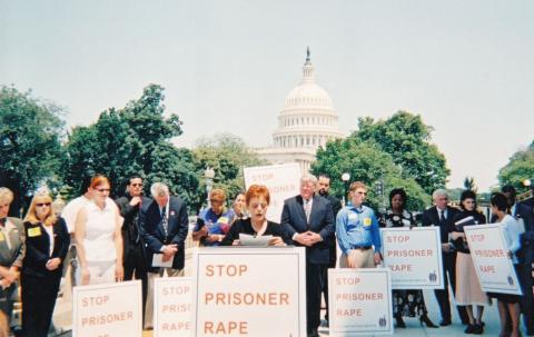 woman standing behind a podium along with several people behind her. There are signs that read "Stop Prisoner Rape." The US Capitol building can be seen in the background.