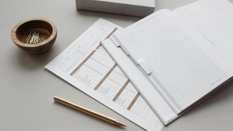 photo of a open notebook on a table with a worksheet beneath it. A pen and paper clips are situated nearby.